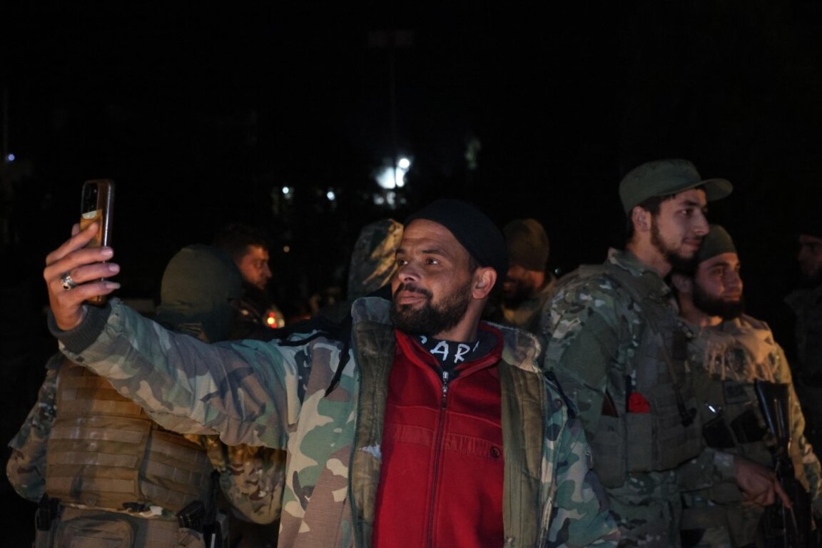 Член на бунтовническа групировка си прави селфи на фона на други бойци,  празнуващи превземането на Дамаск, Хомс, Сирия, сутринта на 8 декември 2024 г.